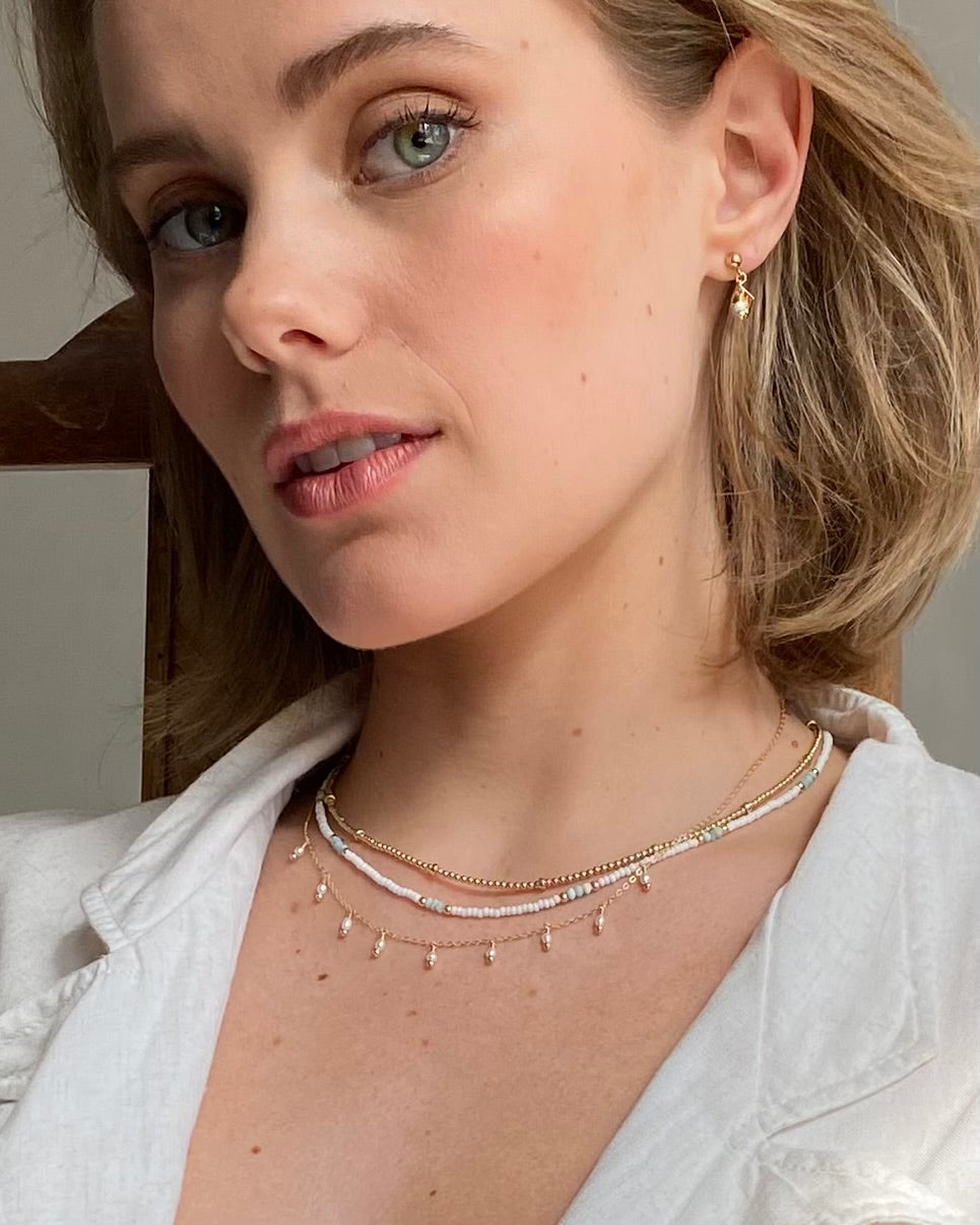 Woman wearing a gold pearl necklaces layered with gold necklaces and pearl earrings with a neutral background