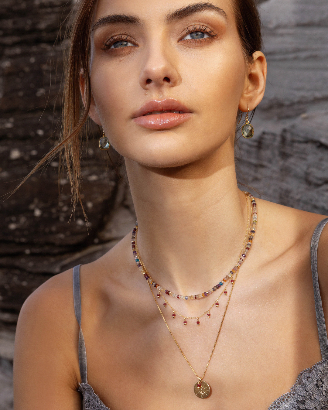 Woman wearing layered gold ruby gemstone necklaces and green amethyst earrings against a stone wall background