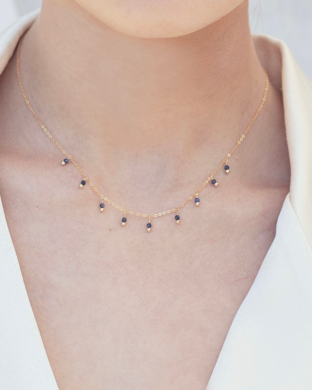 Close up of a woman's neck wearing a cream blazer and a gold sapphire gemstone necklace 