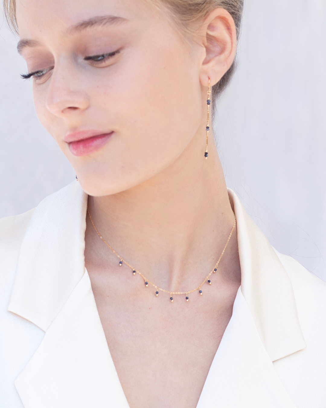 Close-up of a woman in a cream blazer wearing a delicate sapphire necklace and earring against a light background