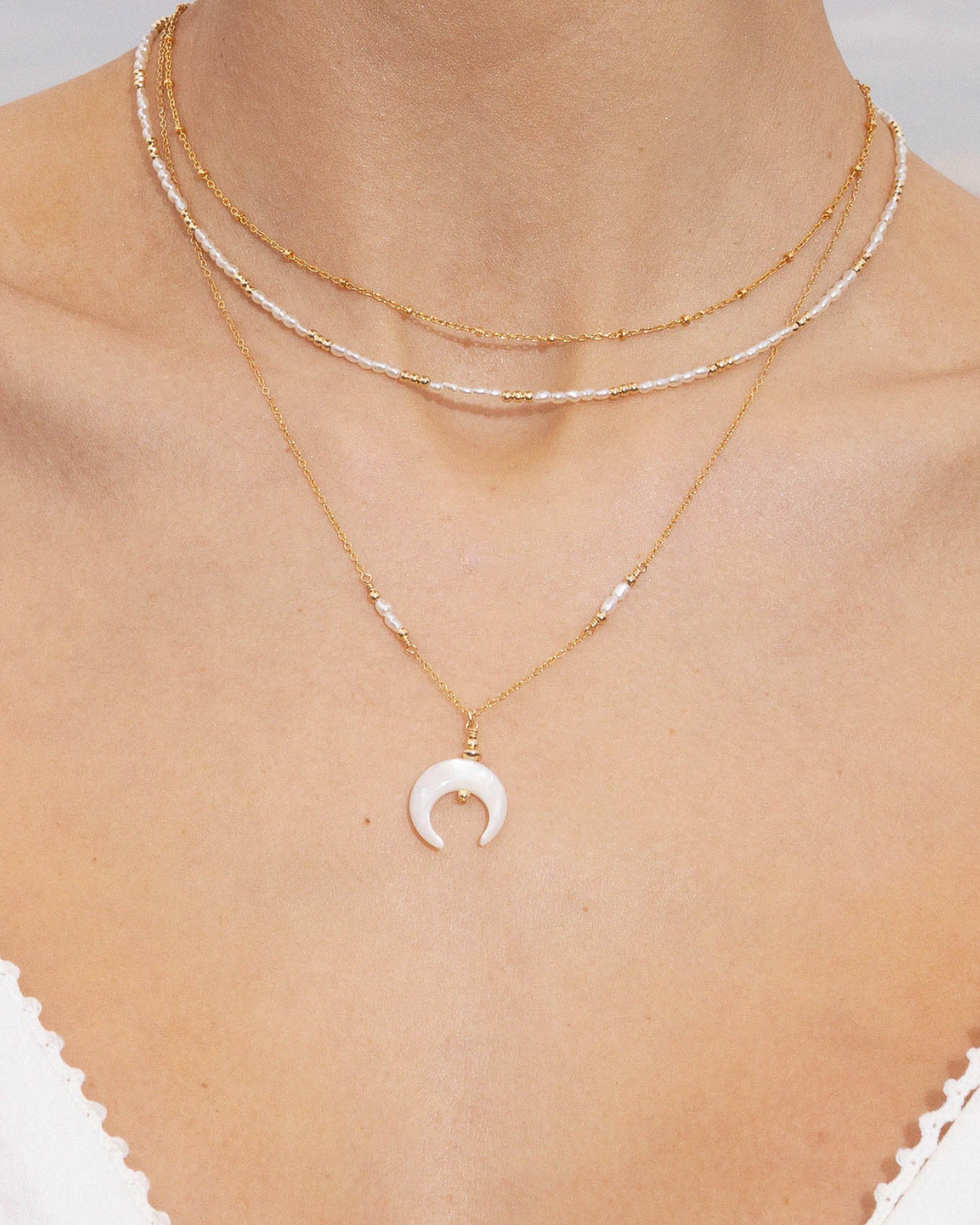 Close up of a woman's neck wearing layered gold pearl necklaces, featuring a necklace with mother of pearl crescent moon pendant