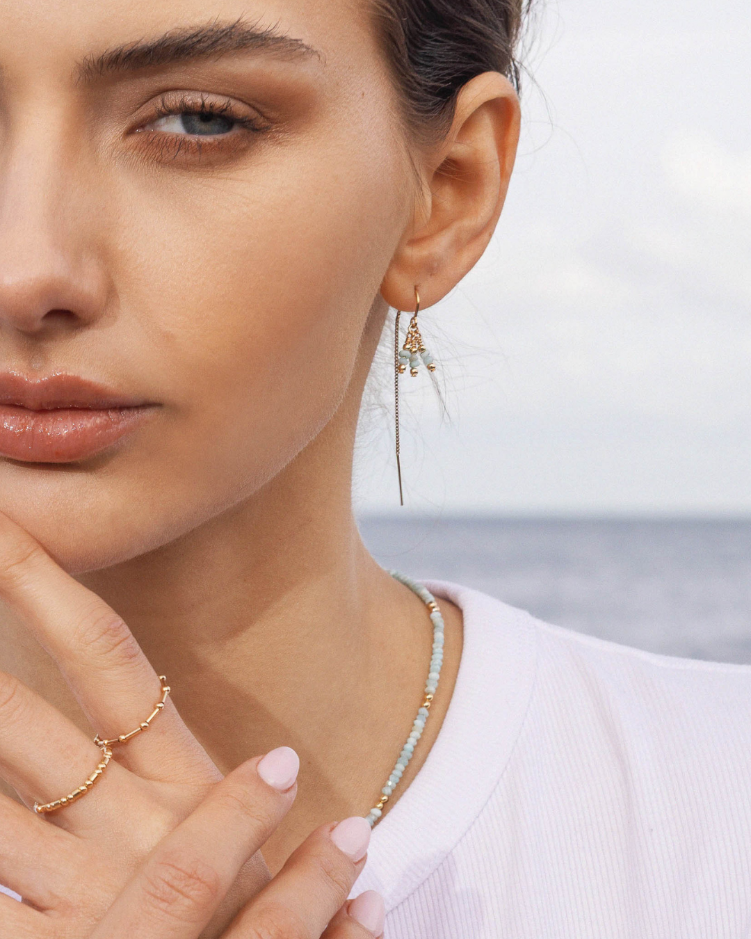 Close-up of a woman wearing a white top with gold beaded rings, gold thread larimar gemstone earring and a matching larimar necklace with a blurred ocean background
