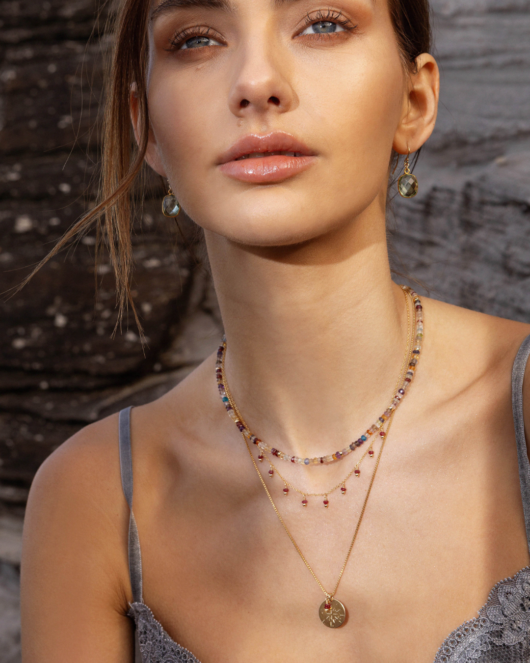 Woman wearing green amethyst gold earrings with a multi-gemstone necklace layered with a ruby necklaces with a natural background