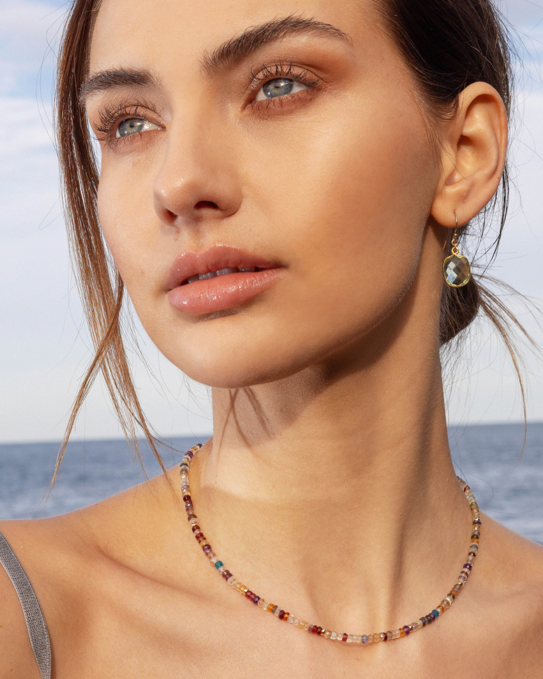 Woman wearing green amethyst gold earrings with a colourful multi gemstone beaded necklace with a blurred ocean background