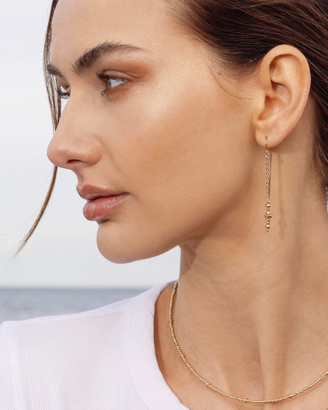 Close-up of a woman wearing gold thread earrings and a gold beaded necklace against a blurred ocean background
