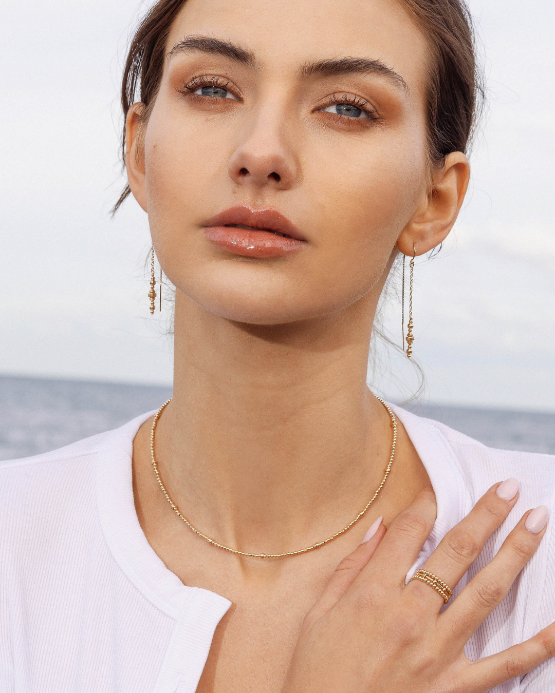 Woman wearing gold jewellery including thread earrings, necklace, and rings with a blurred ocean background