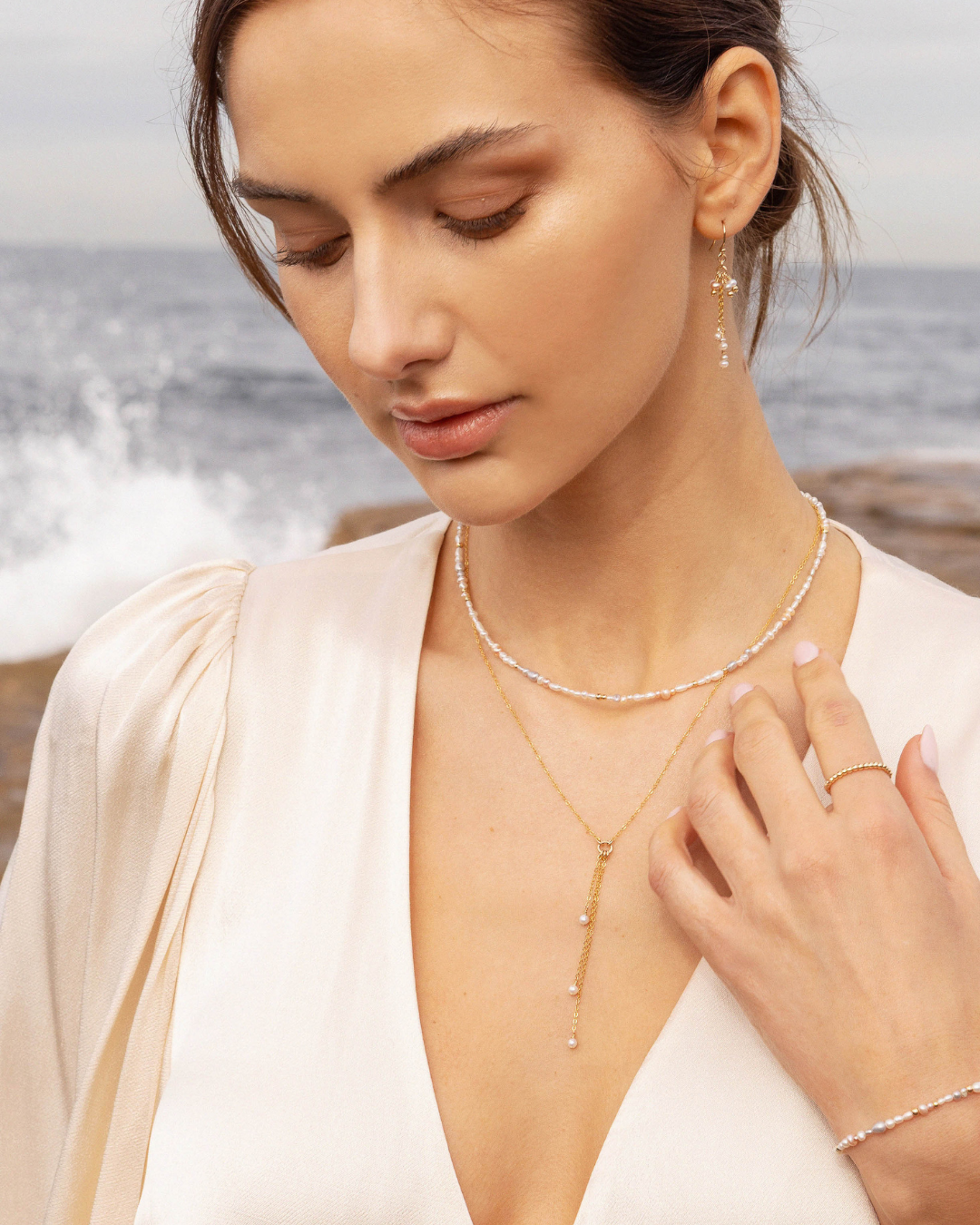 Woman wearing a cream satin top and gold pearl jewellery by the ocean
