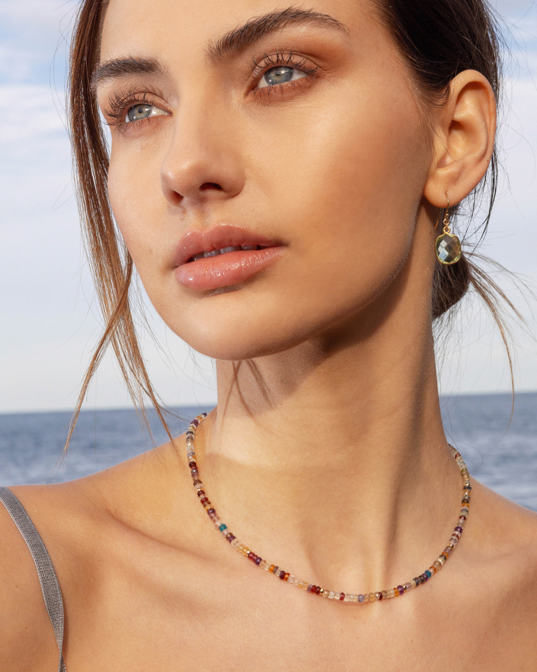 Close up of a woman wearing a gold gemstone beaded necklace with a green amethyst earring with a blurred ocean background
