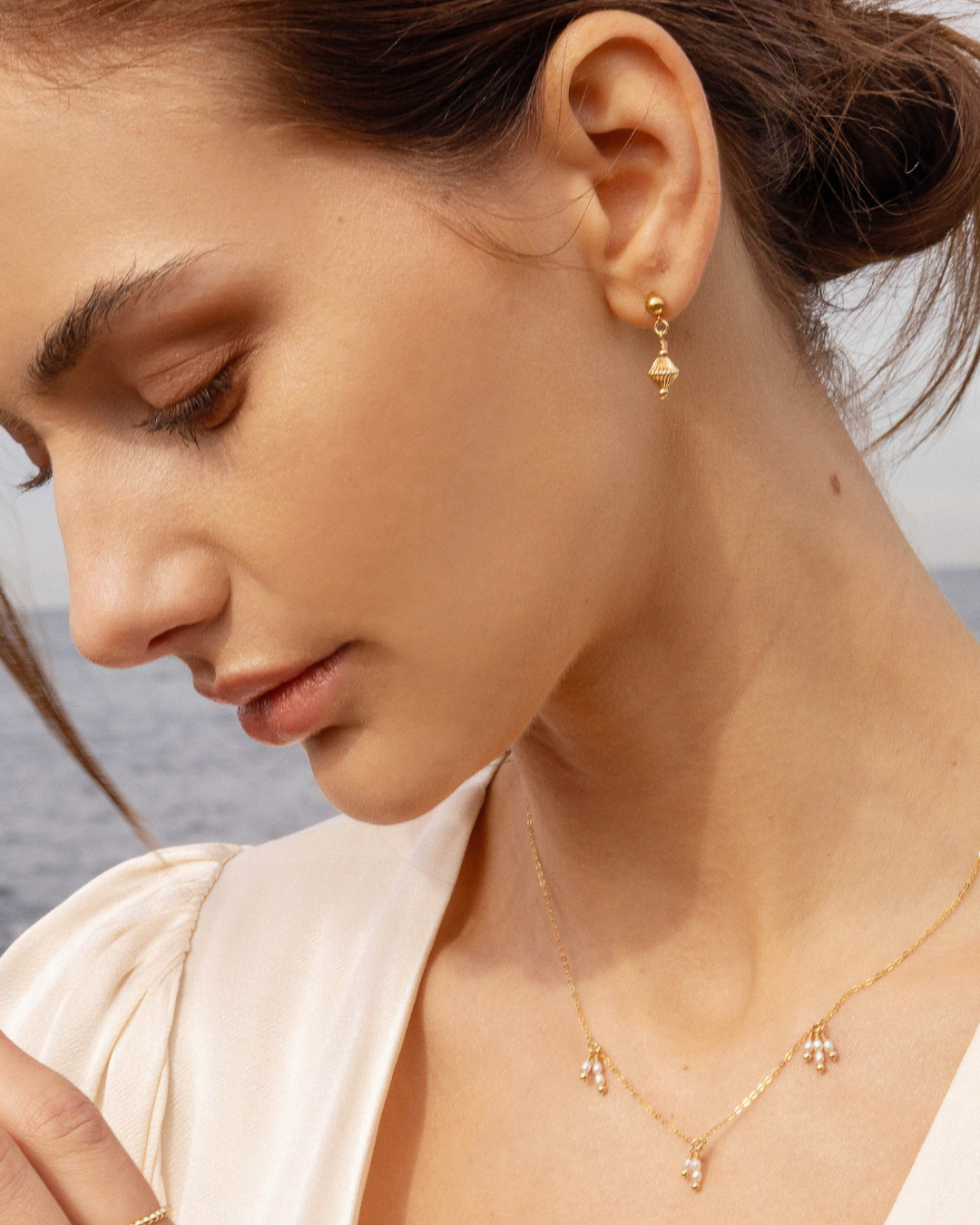 Close-up of a woman wearing gold stud drop earrings and delicate necklace with an ocean background