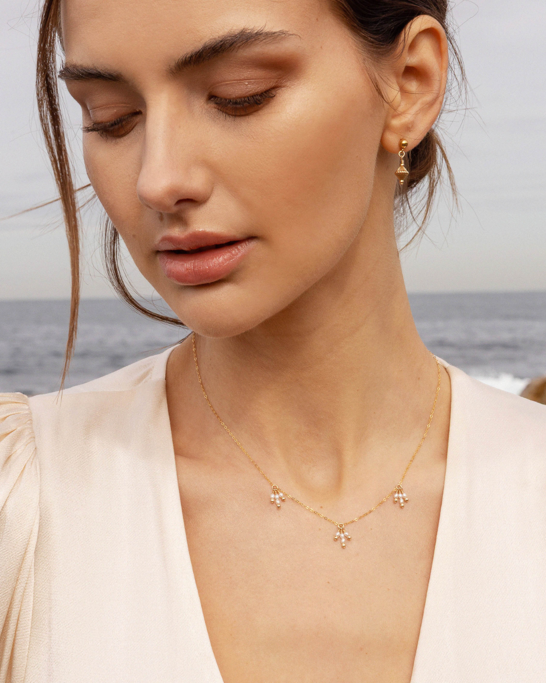 Woman wearing a delicate gold pearl necklace and gold earrings with a blurred ocean background