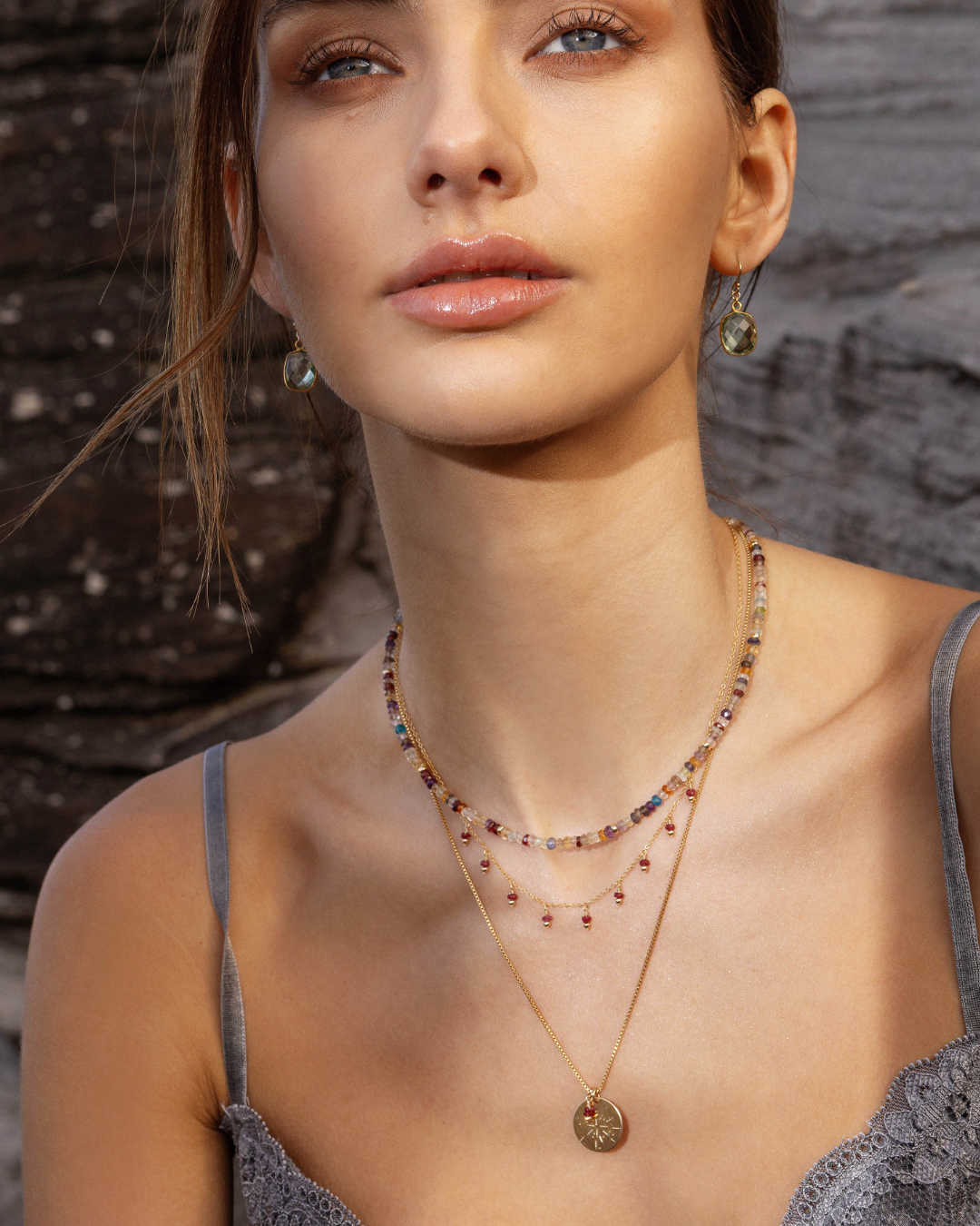 Woman wearing layered necklaces and green amethyst earrings with a stone background