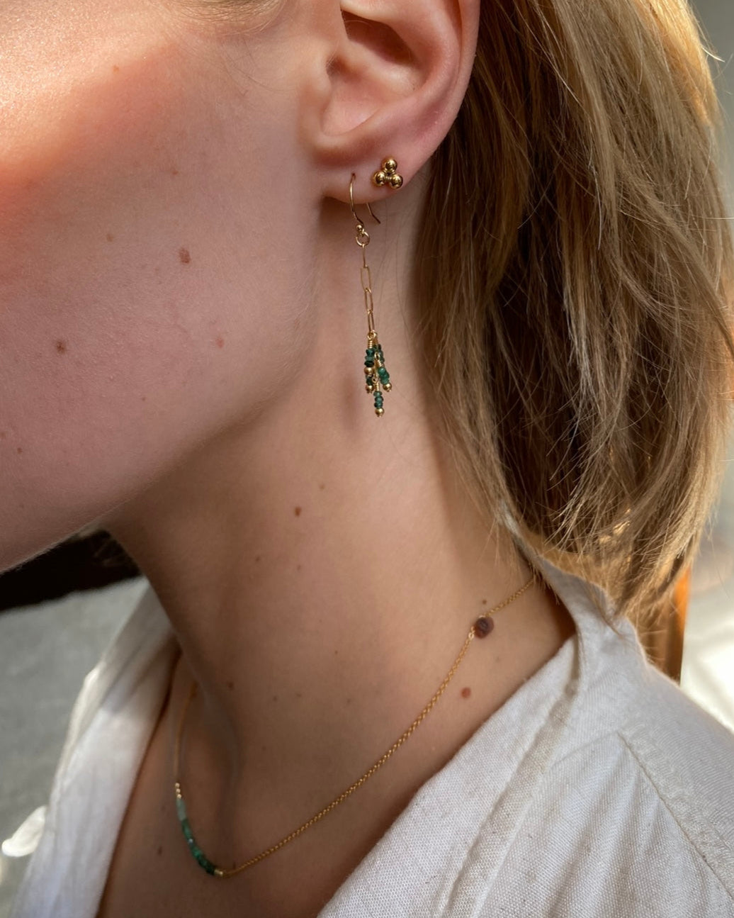 Close-up of a person wearing a gold stud earring and emerald drop earring with a gold emerald necklace. 
