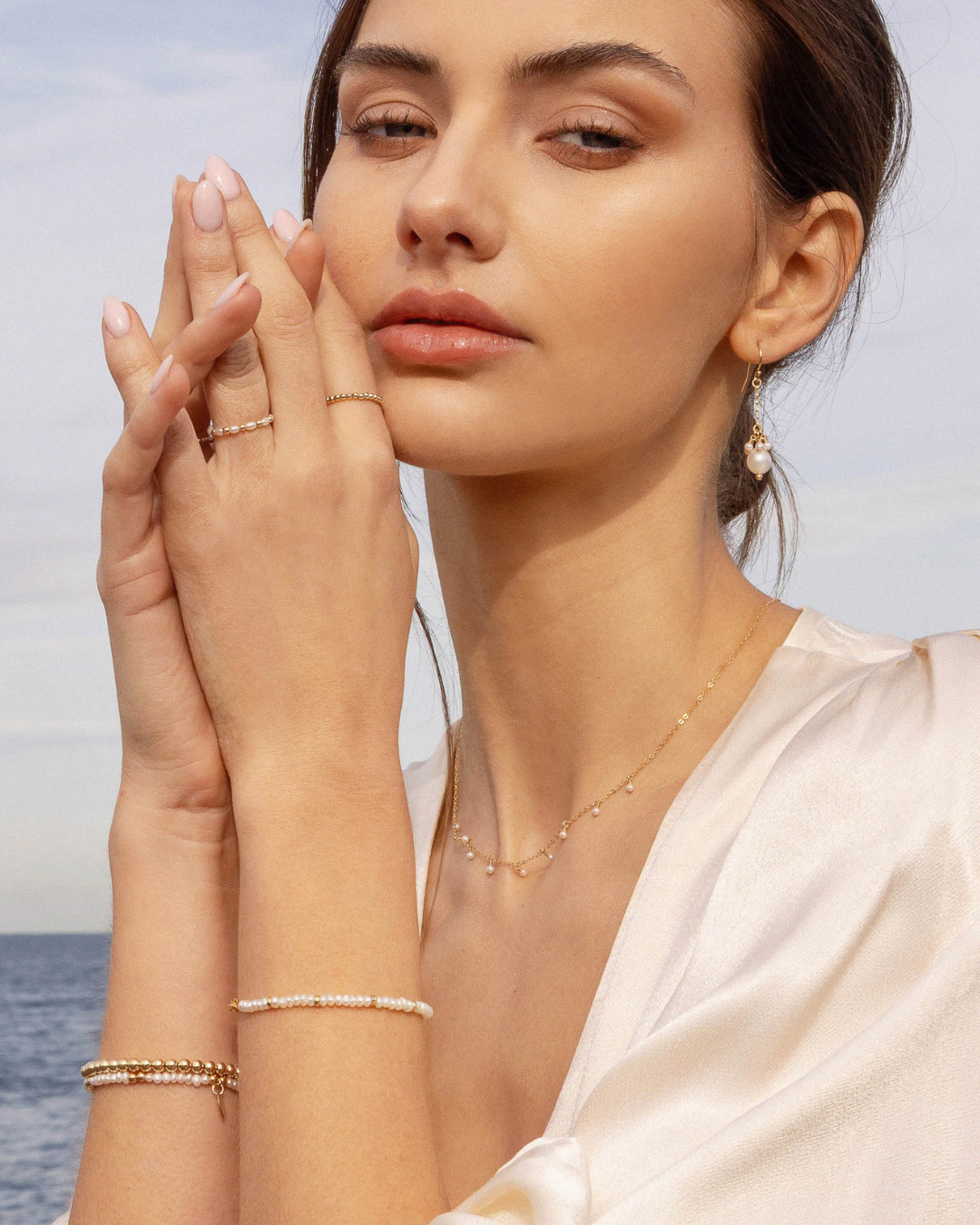 Woman wearing gold pearl drop earrings, pearl bracelets, rings and necklace with an ocean background.