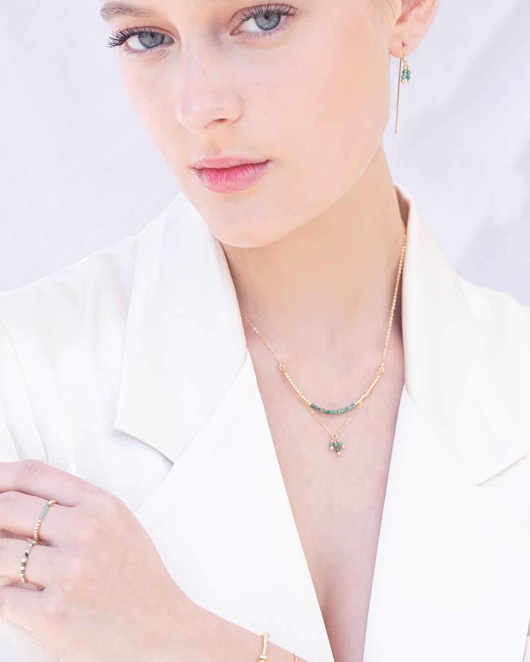 Close-up of a woman wearing a cream blazer, emerald necklace, thread earrings and rings with a white background
