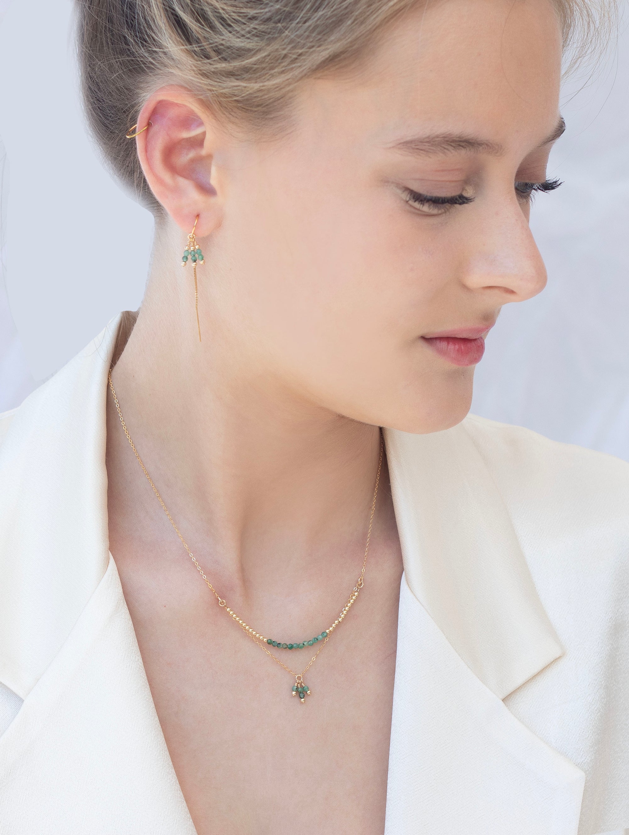 Woman wearing a gold necklace with emerald gemstones necklace and thread earring with a light background