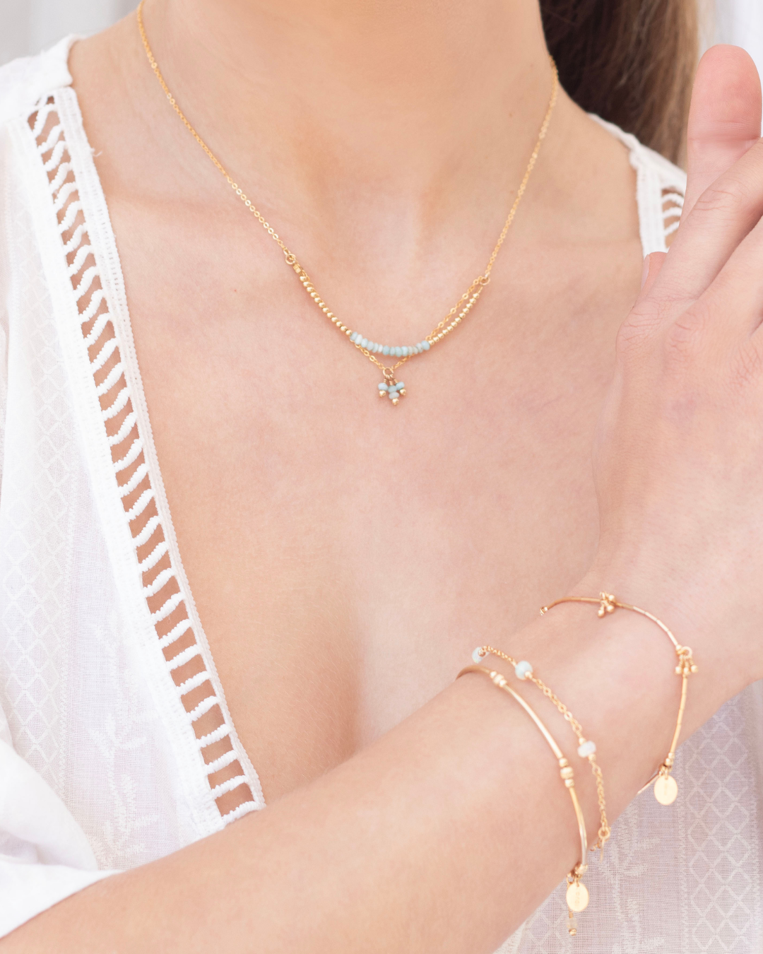Close-up of a woman wearing a gold larimar gemstone necklace and gold beaded bracelets on a white background