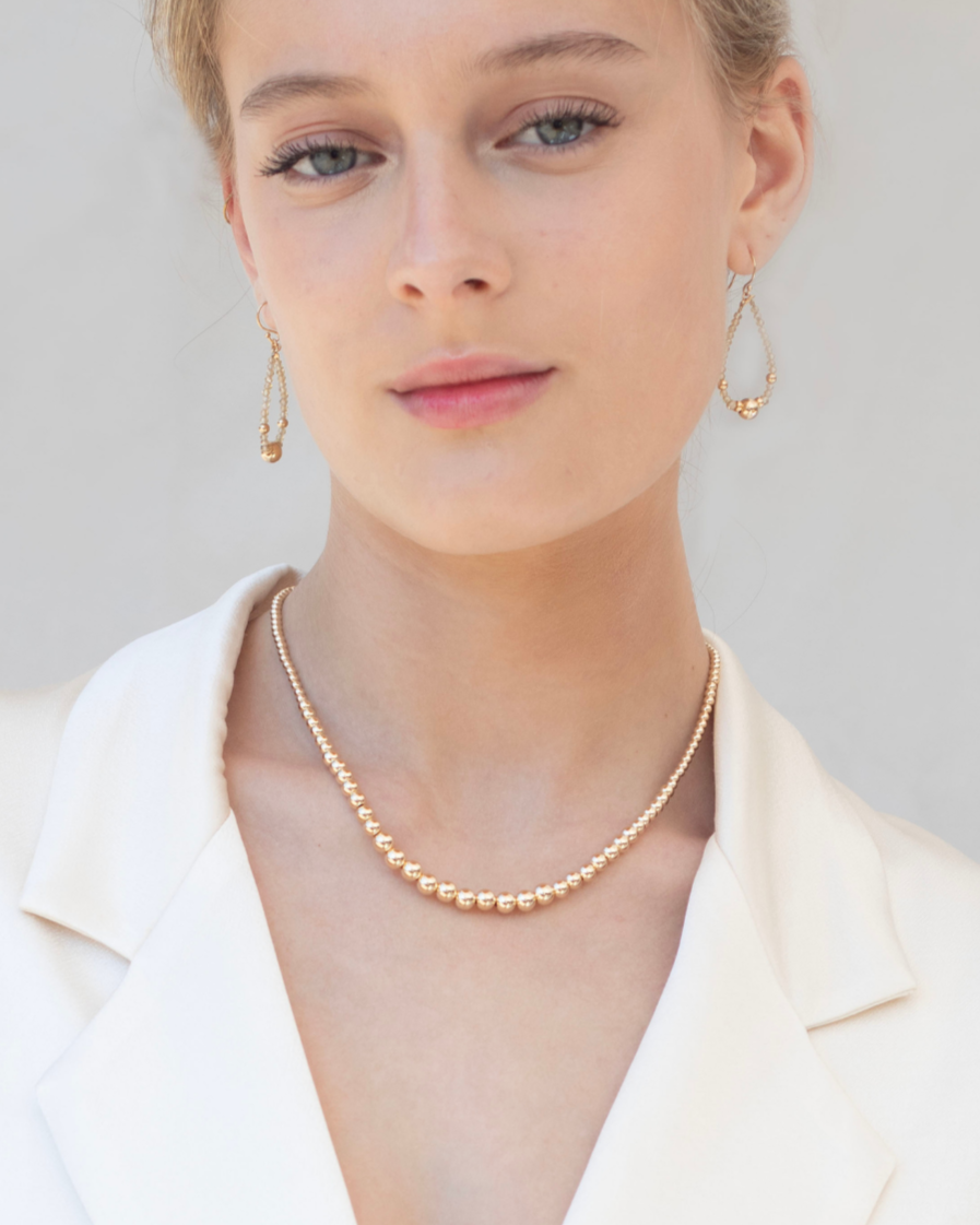 Woman wearing gold and zircon gemstone earrings and gold necklace, wearing a cream satin blazer with a blurred background.