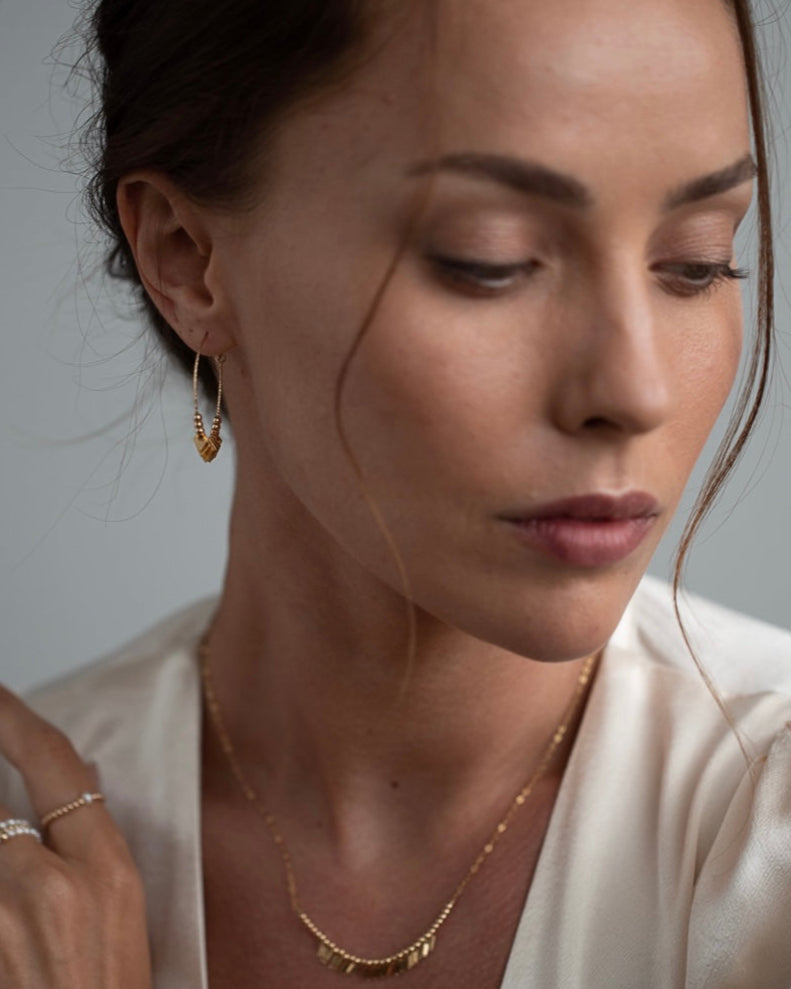 Close-up of a woman wearing gold hoop earrings and a necklace against a neutral background