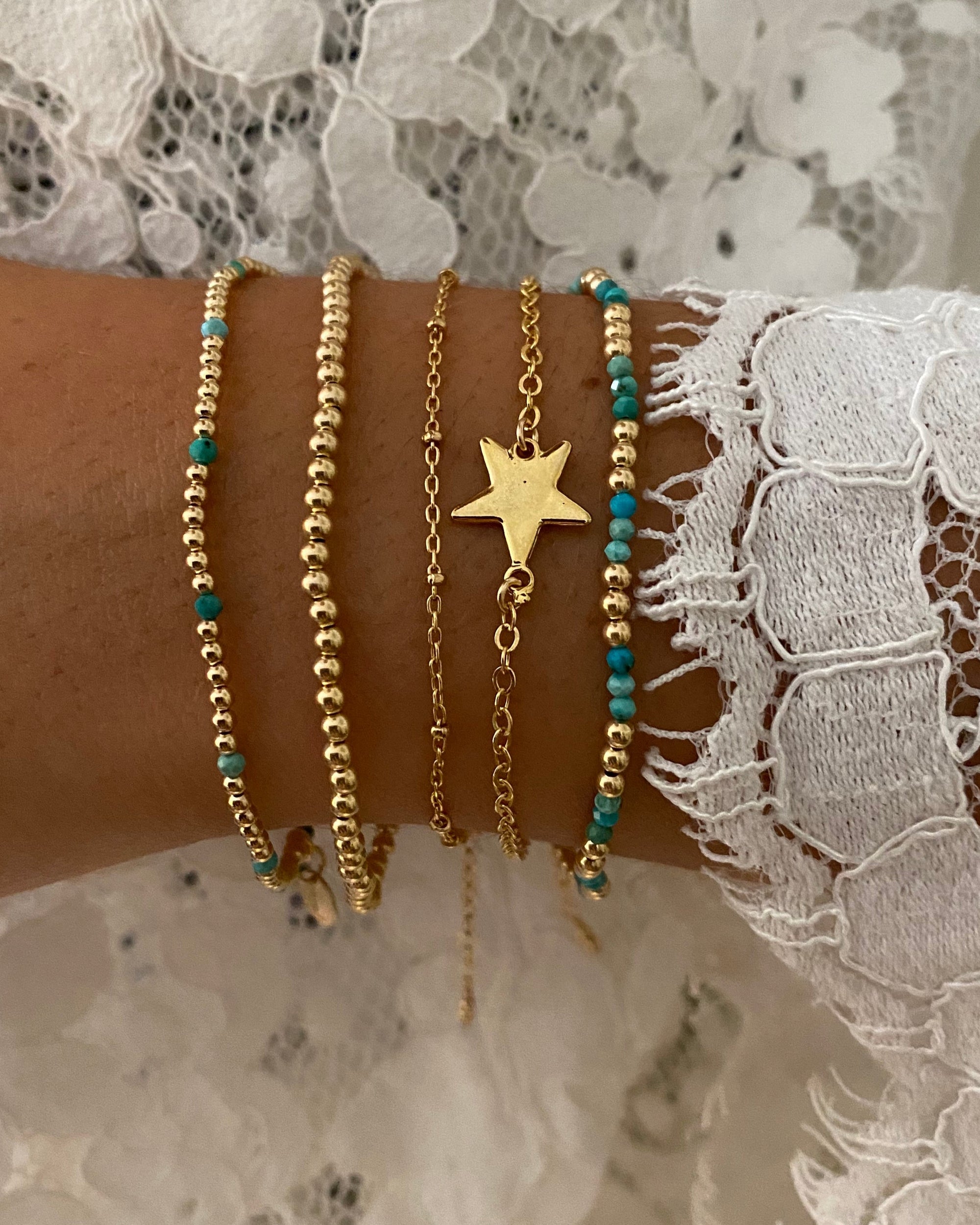 Hand wearing multiple gold and turquoise bracelets with a star charm on a lace background