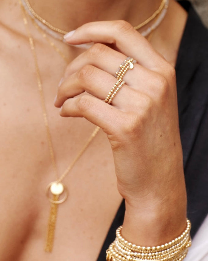 Close-up of a hand wearing gold jewellery including a labradorite ring, gold bracelets, and necklaces.