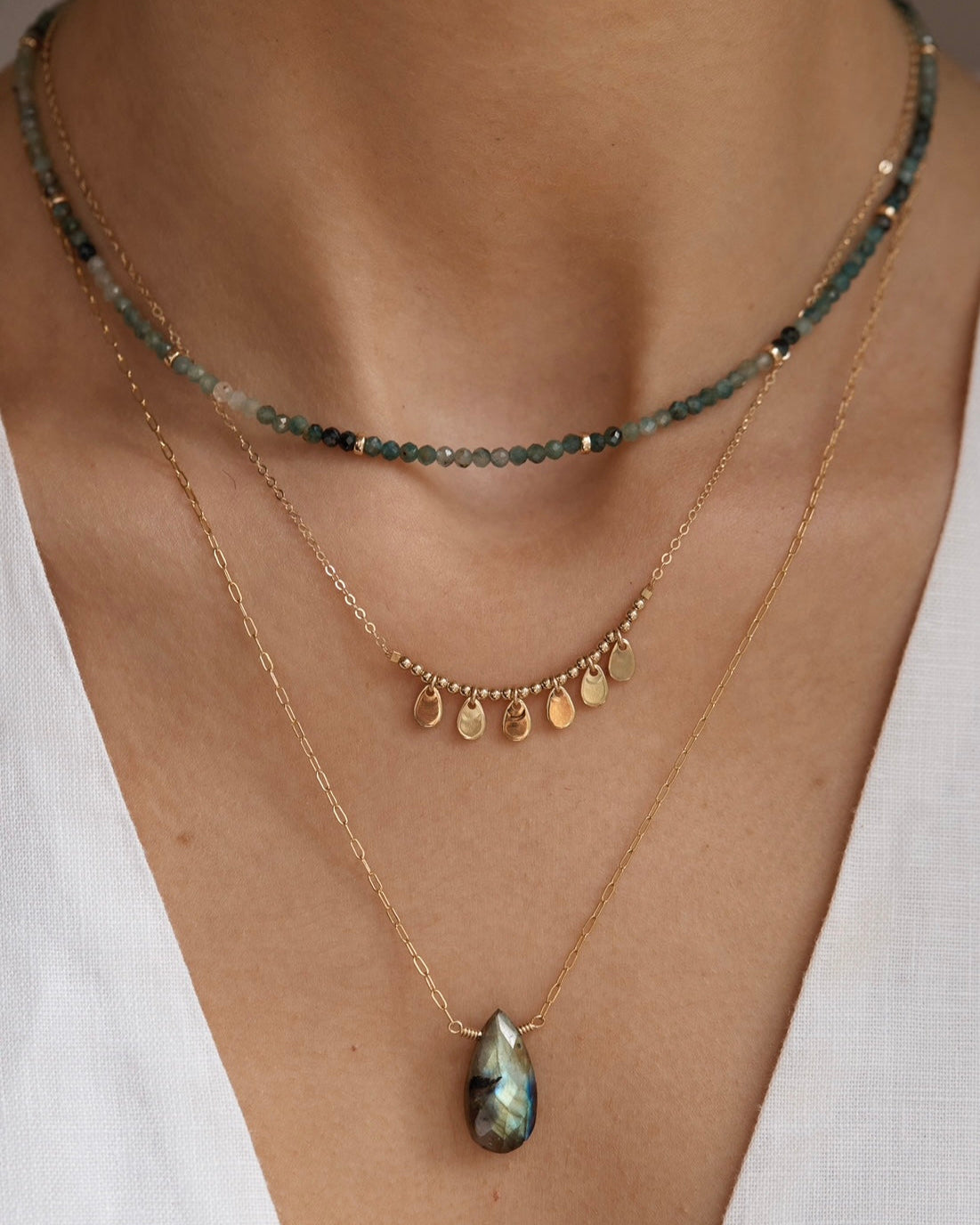Close up photo of three layered gold necklaces featuring grandidierite and labradorite gemstones on a person wearing a white top