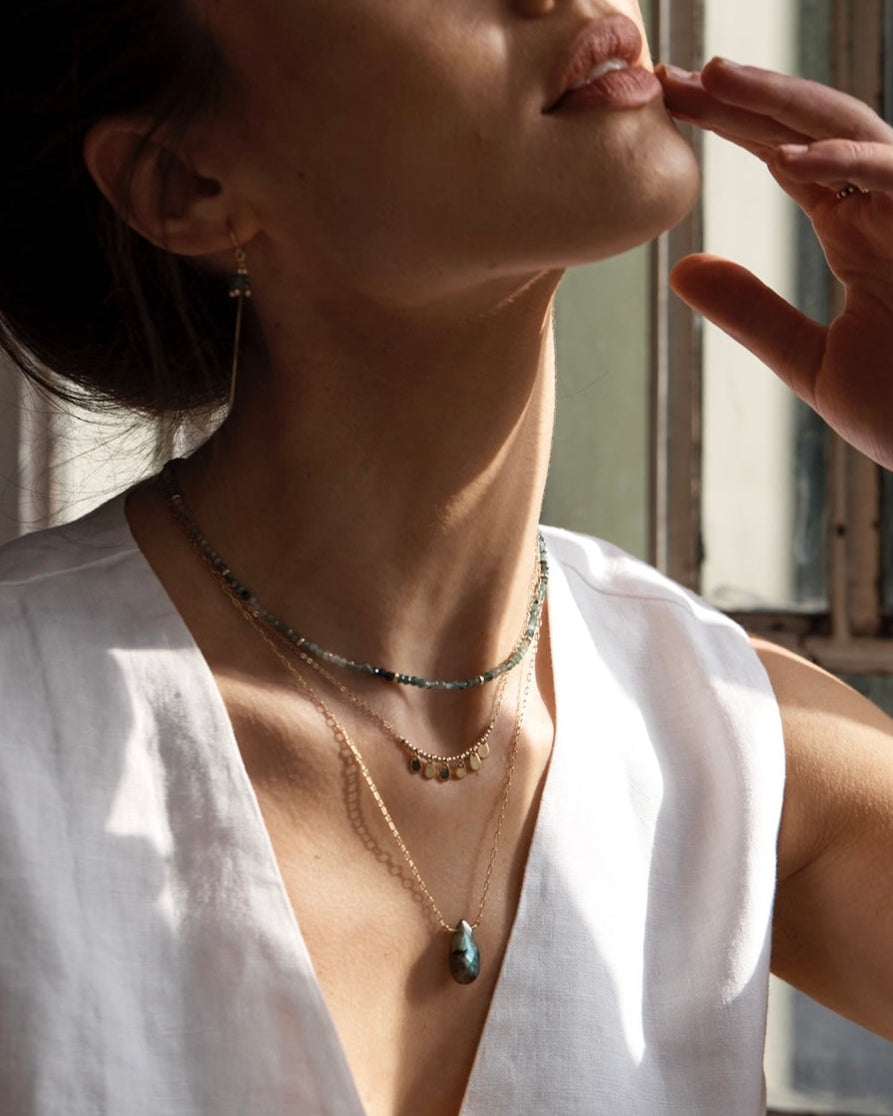 Woman wearing gold layered necklaces with a labradorite pendant, grandidierite gemstones and a delicate charm necklace.