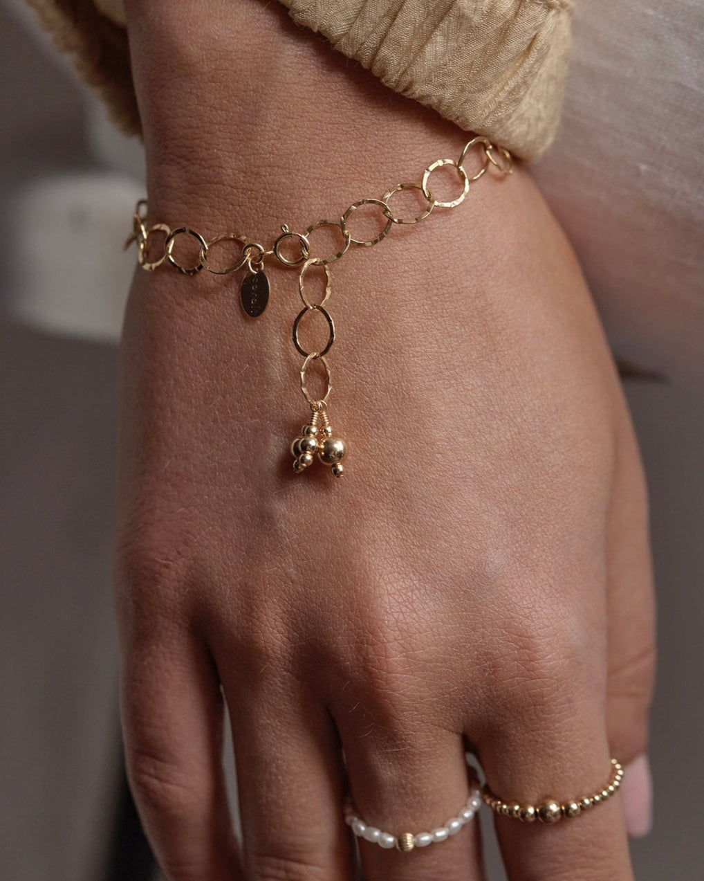 Gold rings and bracelet with gold bead charms on a wrist against a neutral background