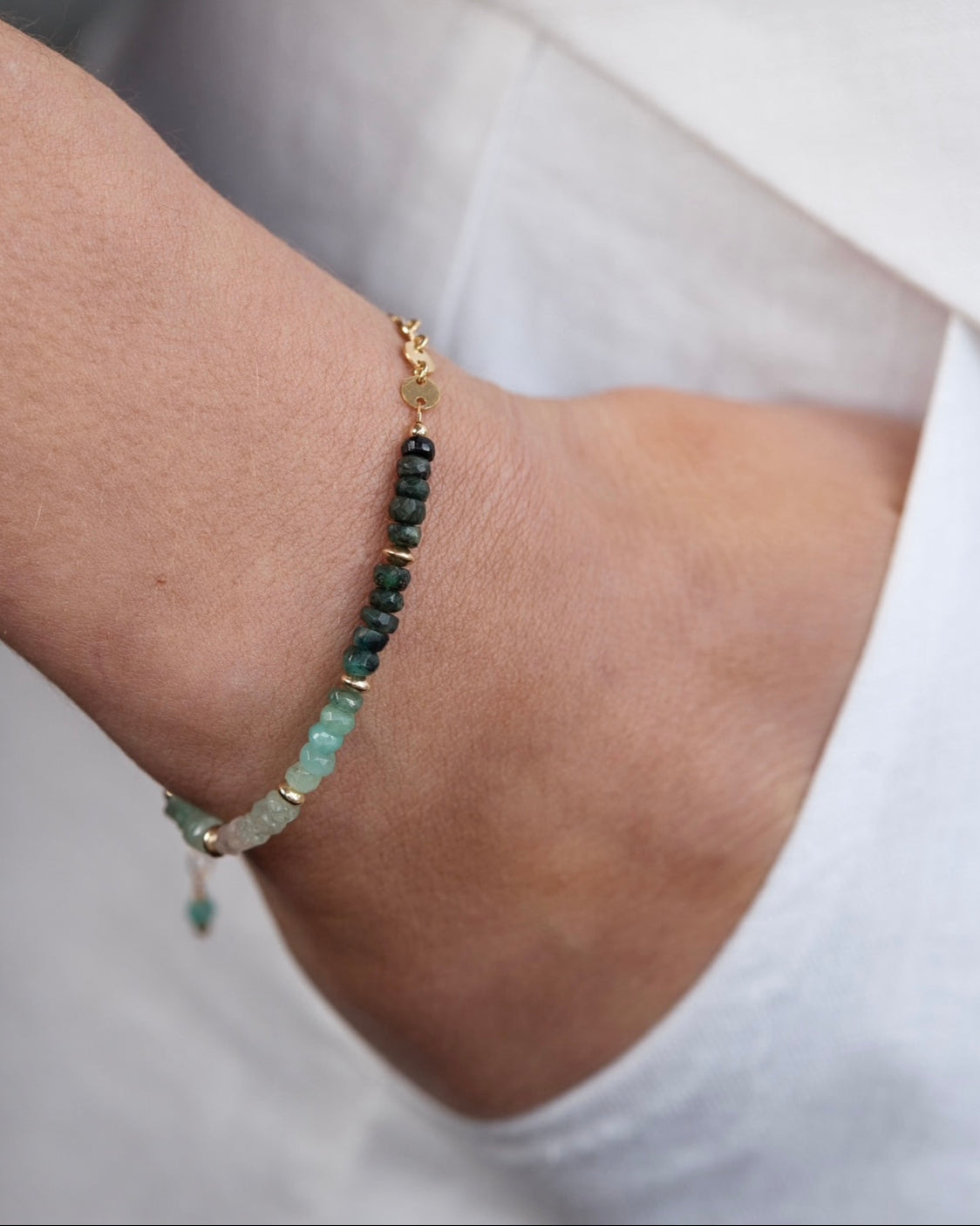 Woman's wrist wearing a gold emerald gemstone bracelet with white linen trousers in the background