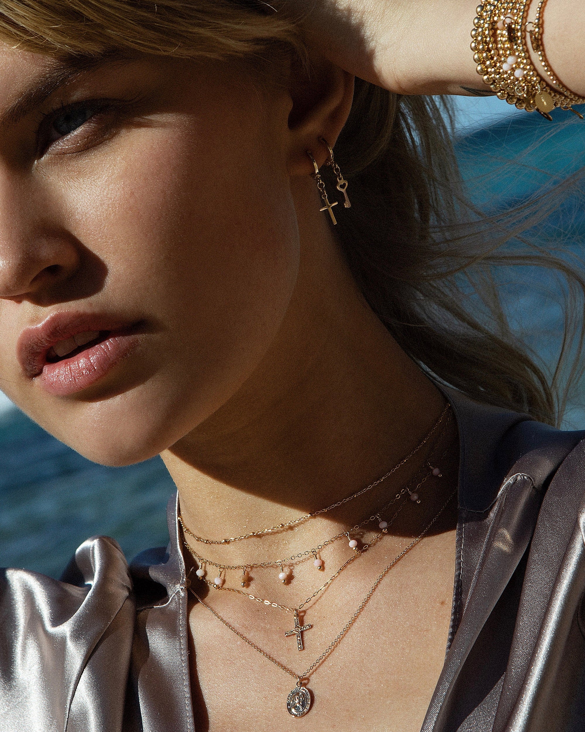 Close-up of a woman wearing gold jewellery including gold pink peruvian opal gemstones bracelets, necklace and hoop earrings with an ocean background