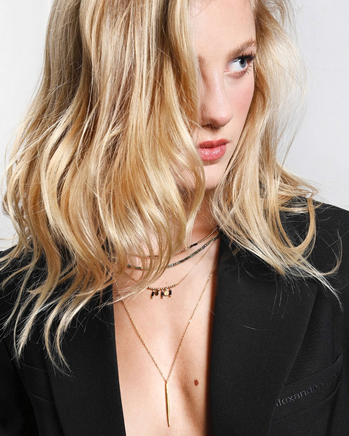 Woman with blonde hair wearing a black blazer and layered gold necklaces with black gemstones against a neutral background
