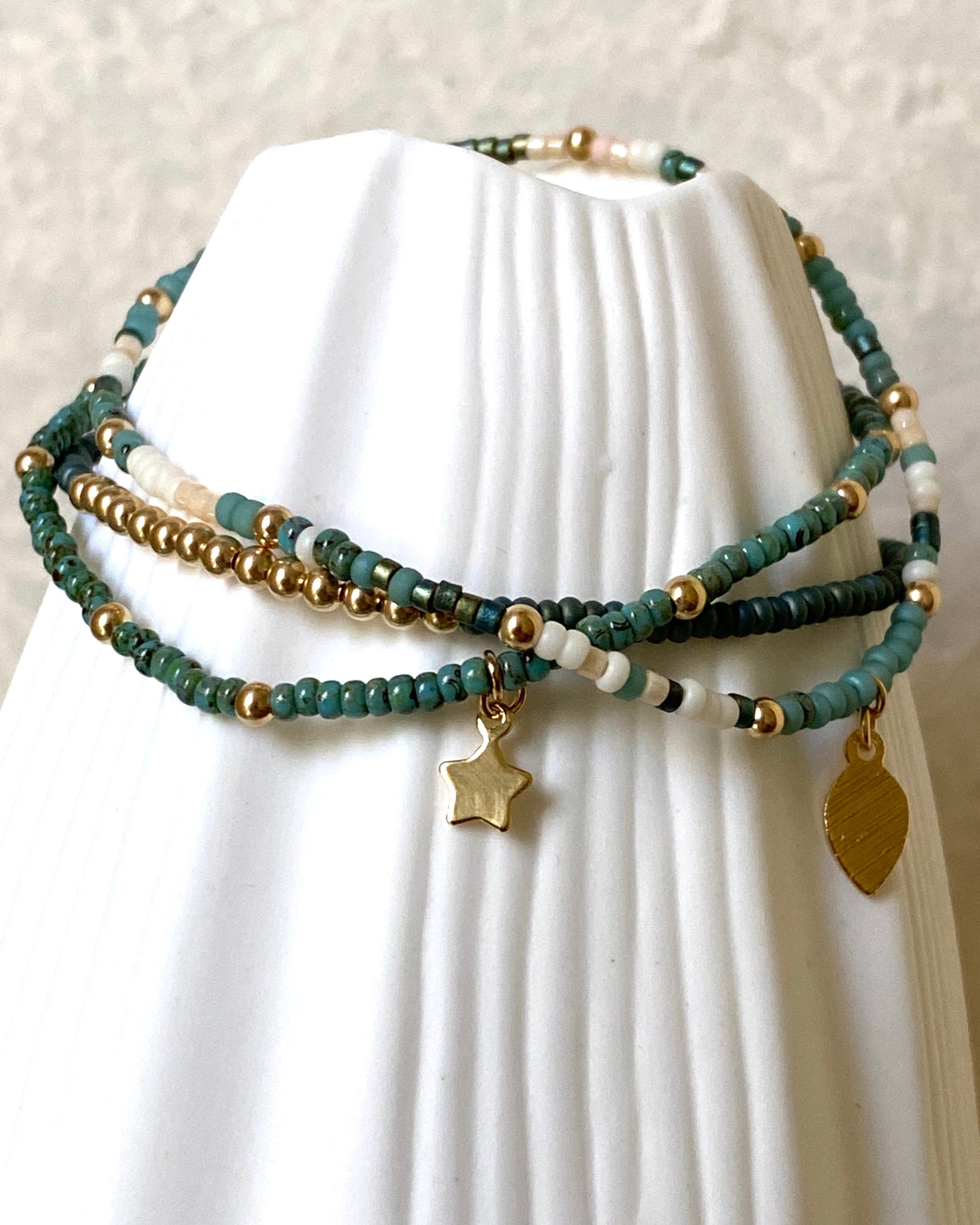Stack of gold and glass beaded bracelets with star and leaf charms displayed on a white vase