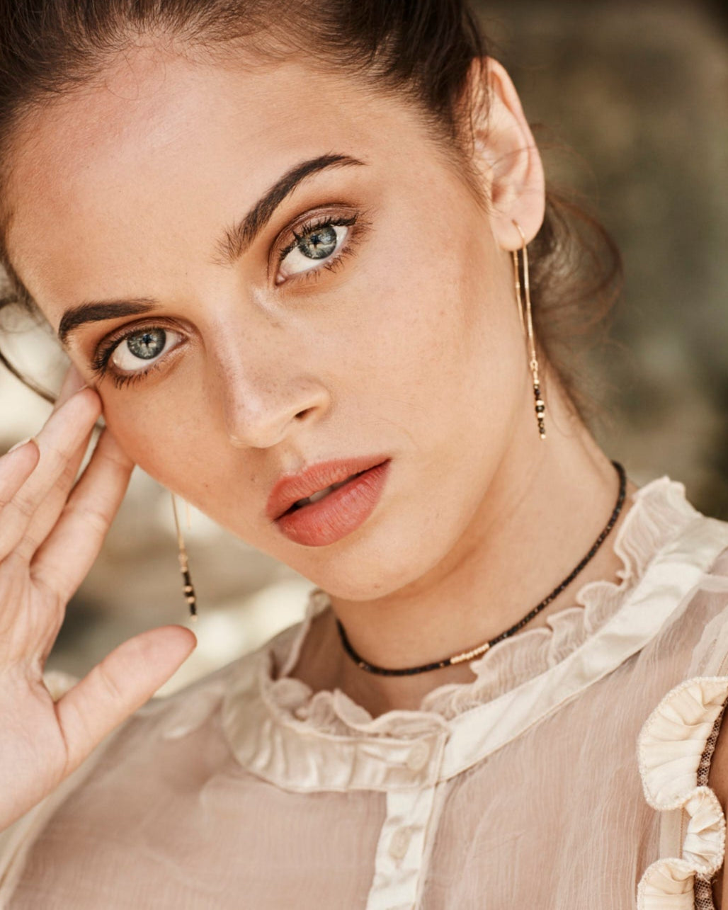 Close-up of a woman wearing gold necklace and earrings with black gemstones and a beige blouse with ruffled details.