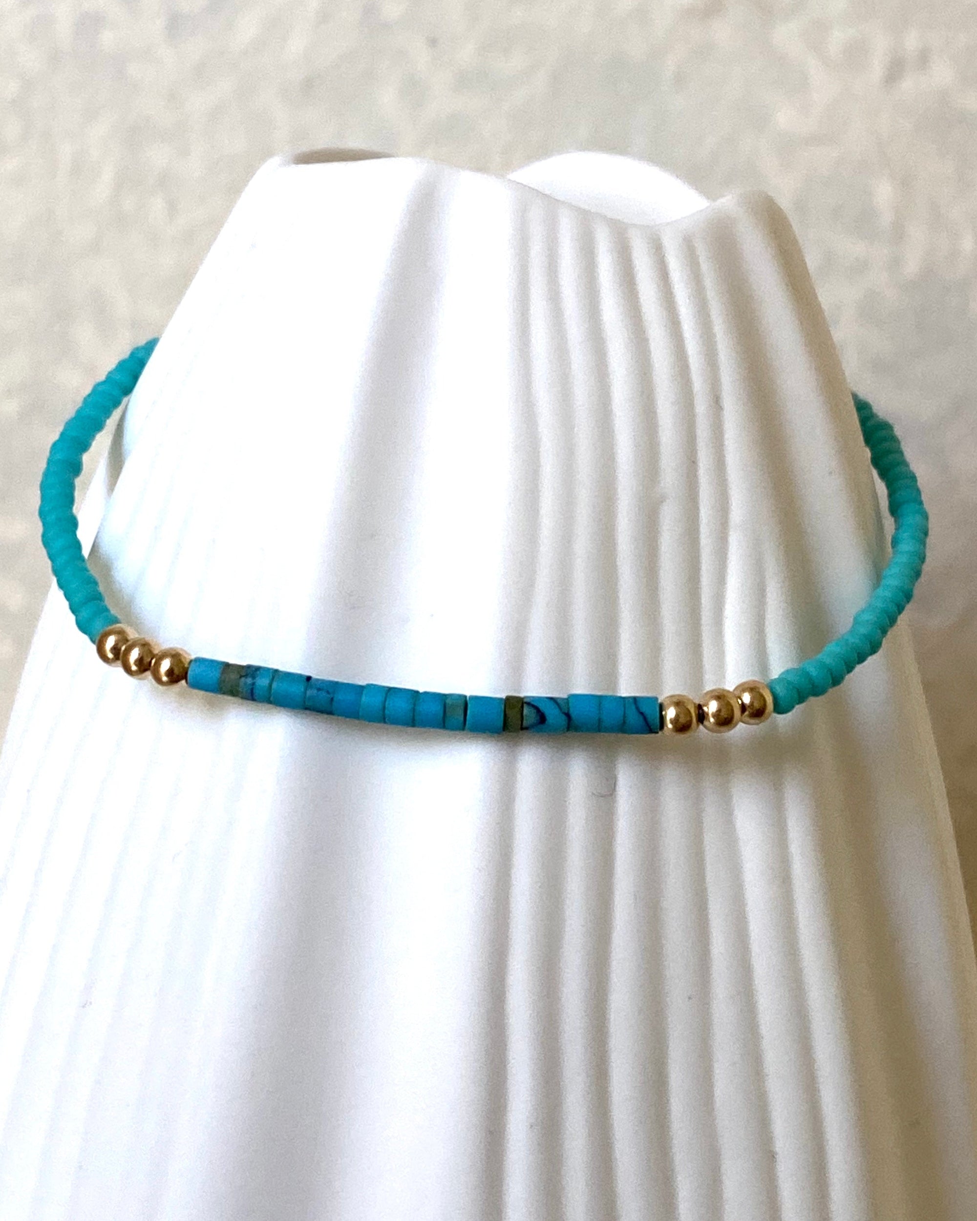Turquoise glass and gold beaded bracelet displayed on a white vase